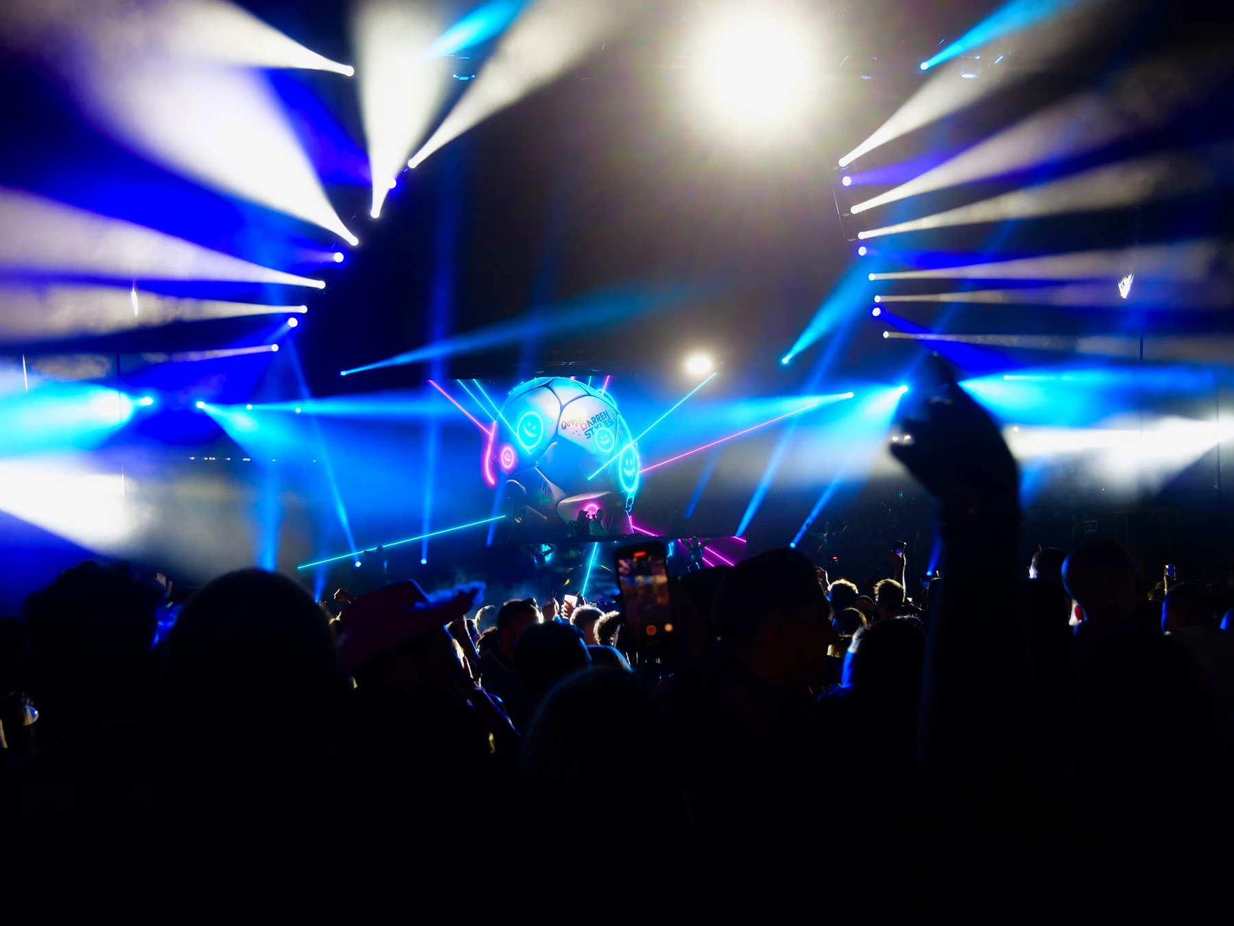 Blue light beams and lasers fanning out from a stage with a giant smiley-faced football set, the crowd filming on phones