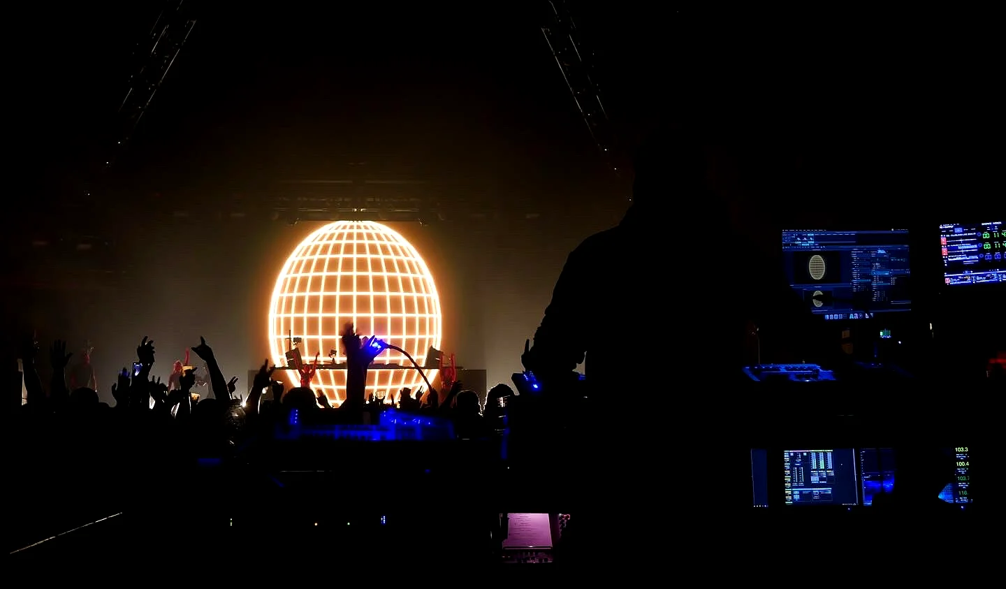A giant glowing disco ball on stage behind the DJ booth, with the crowd's hands raised in silhouette