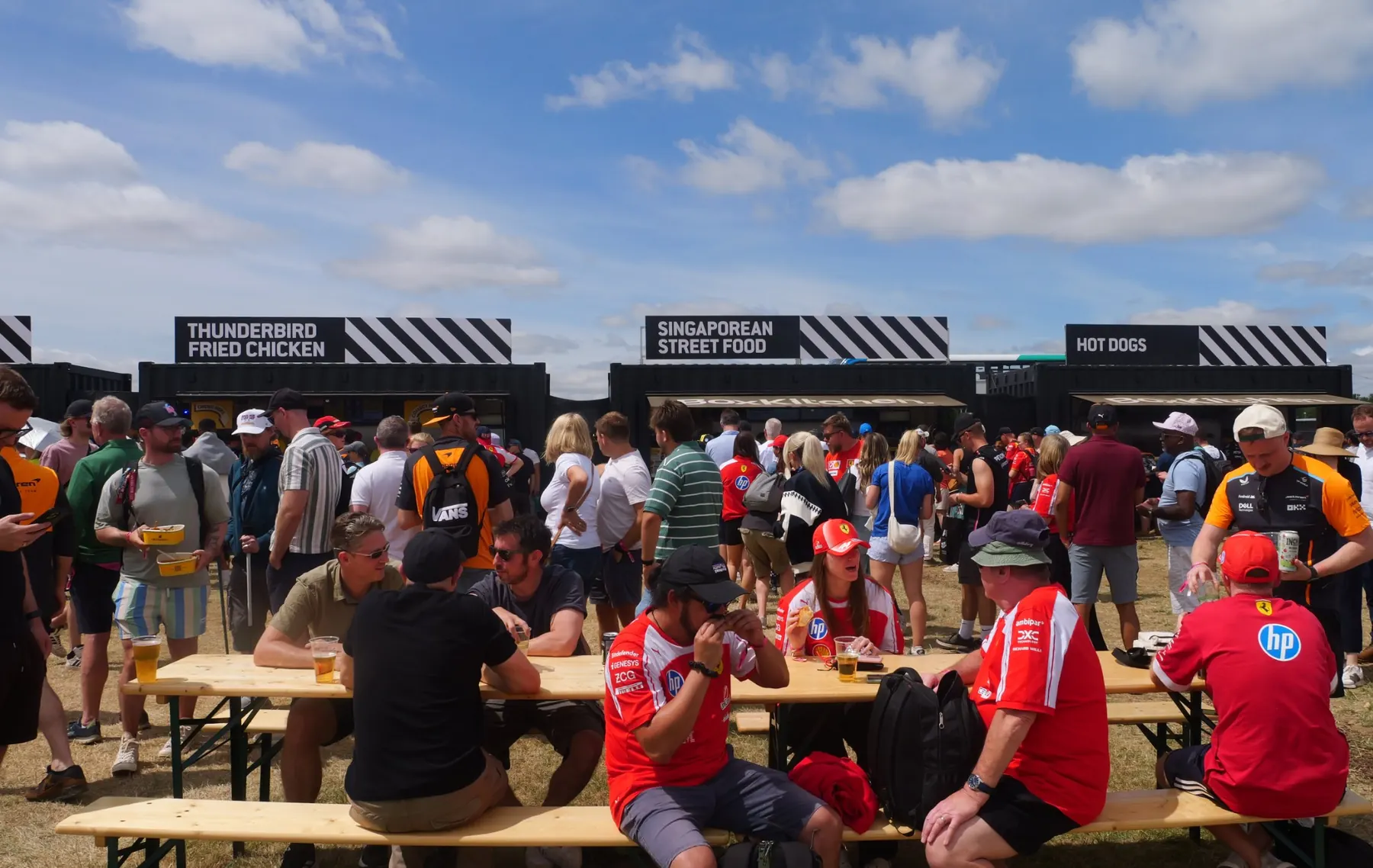 Formula 1 fans eating at benches in front of black BoxKitchen container kitchens at Silverstone during the British Grand Prix