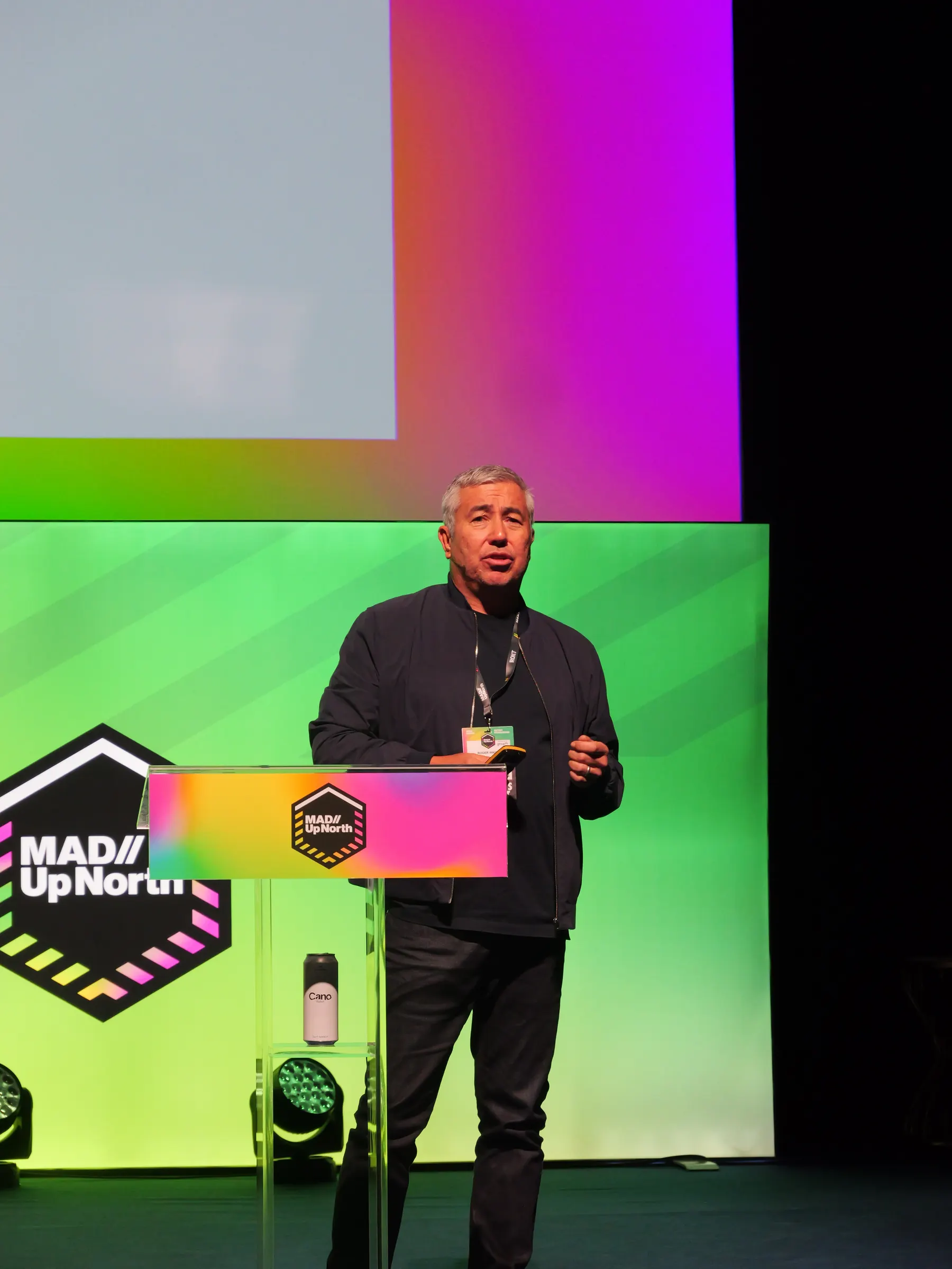 Roger Wade, founder of BOXPARK and now behind BoxKitchen, speaking at a lectern on the pink and green stage at MAD//Fest Up North in Manchester