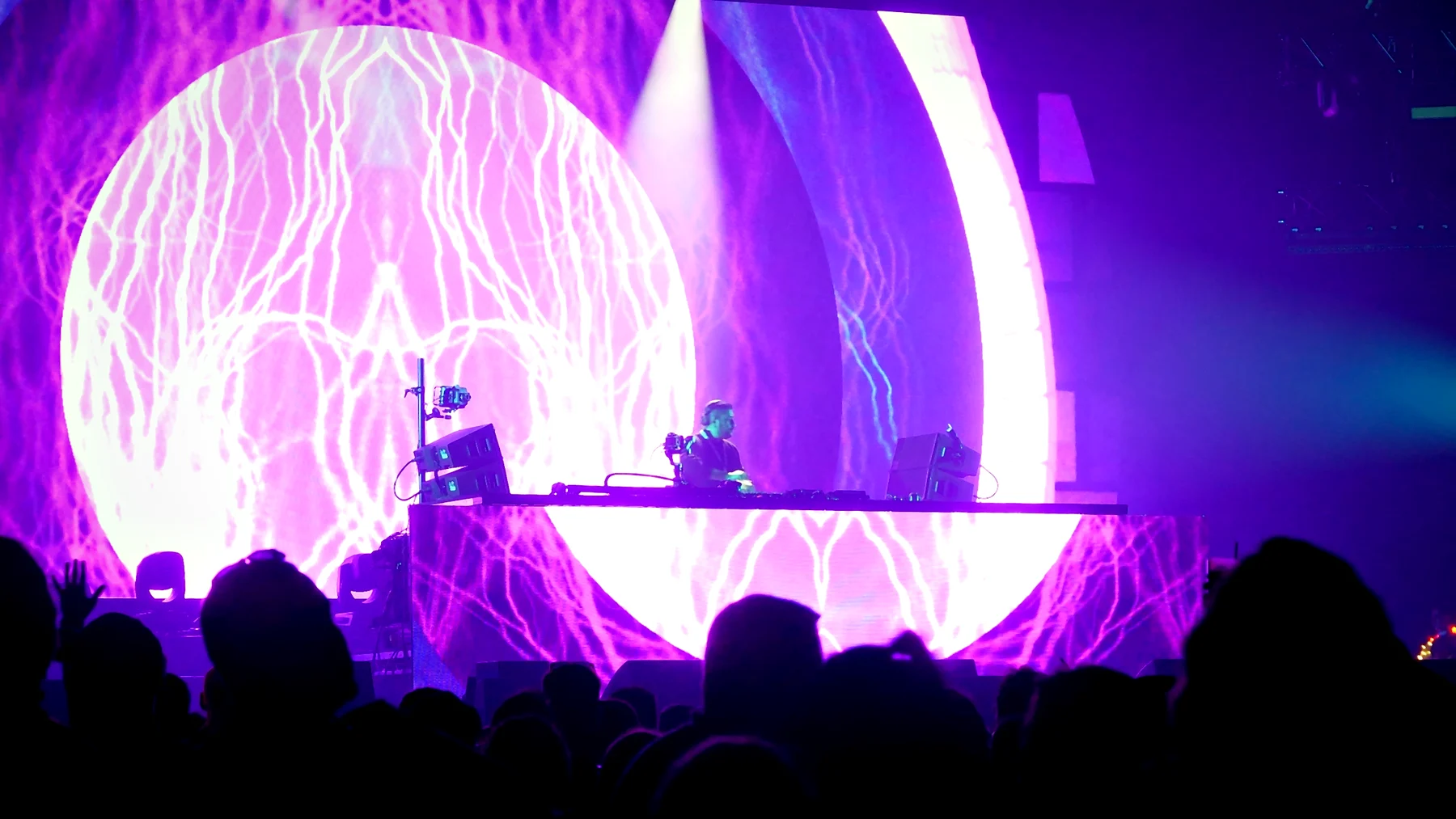 DJ behind a lit booth in front of a giant pink and violet orb of visuals, with the crowd in silhouette