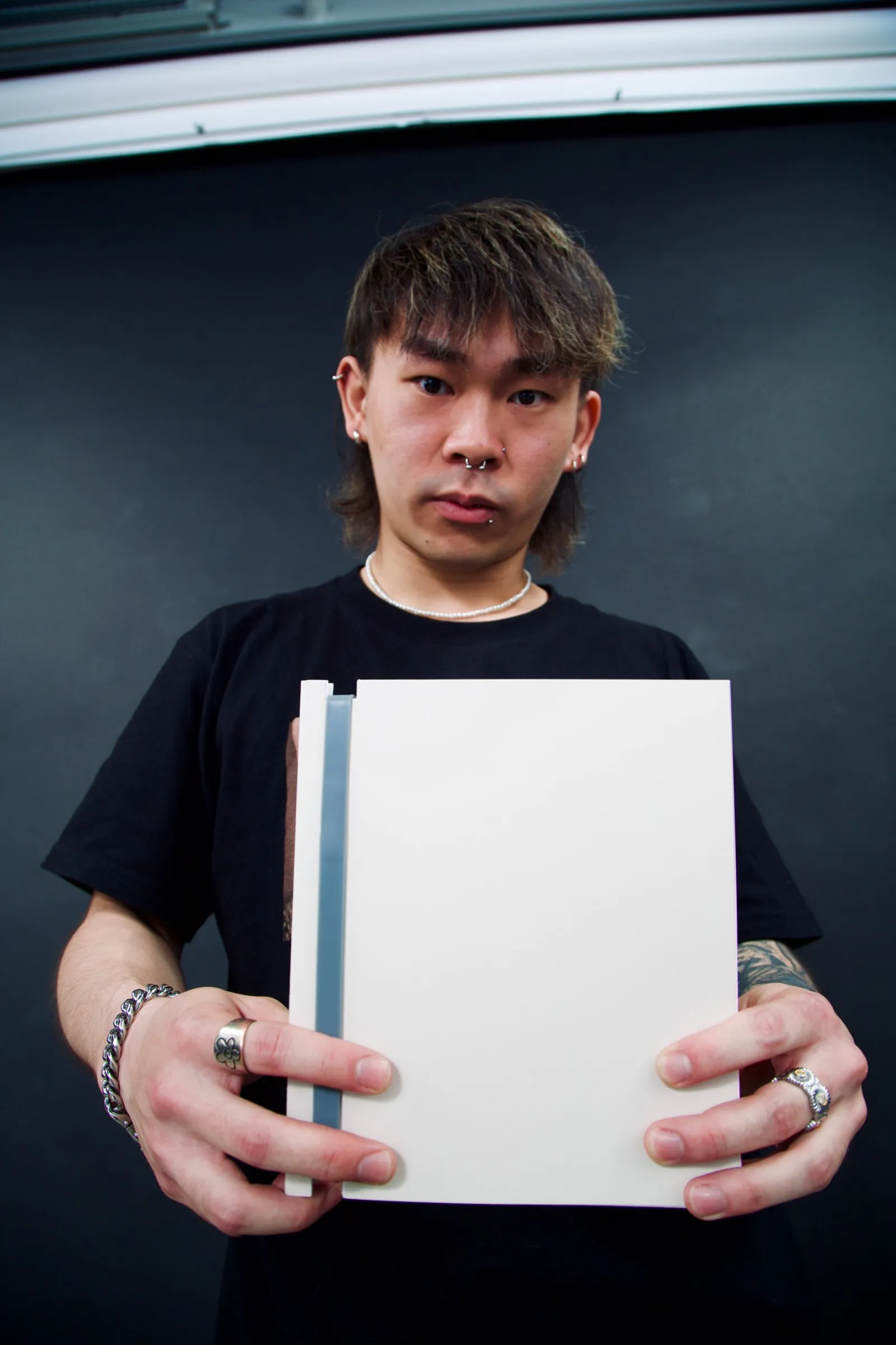 Model in a black t-shirt holding a cream Book for Life refillable notebook with a grey spine, studio portrait against a black wall