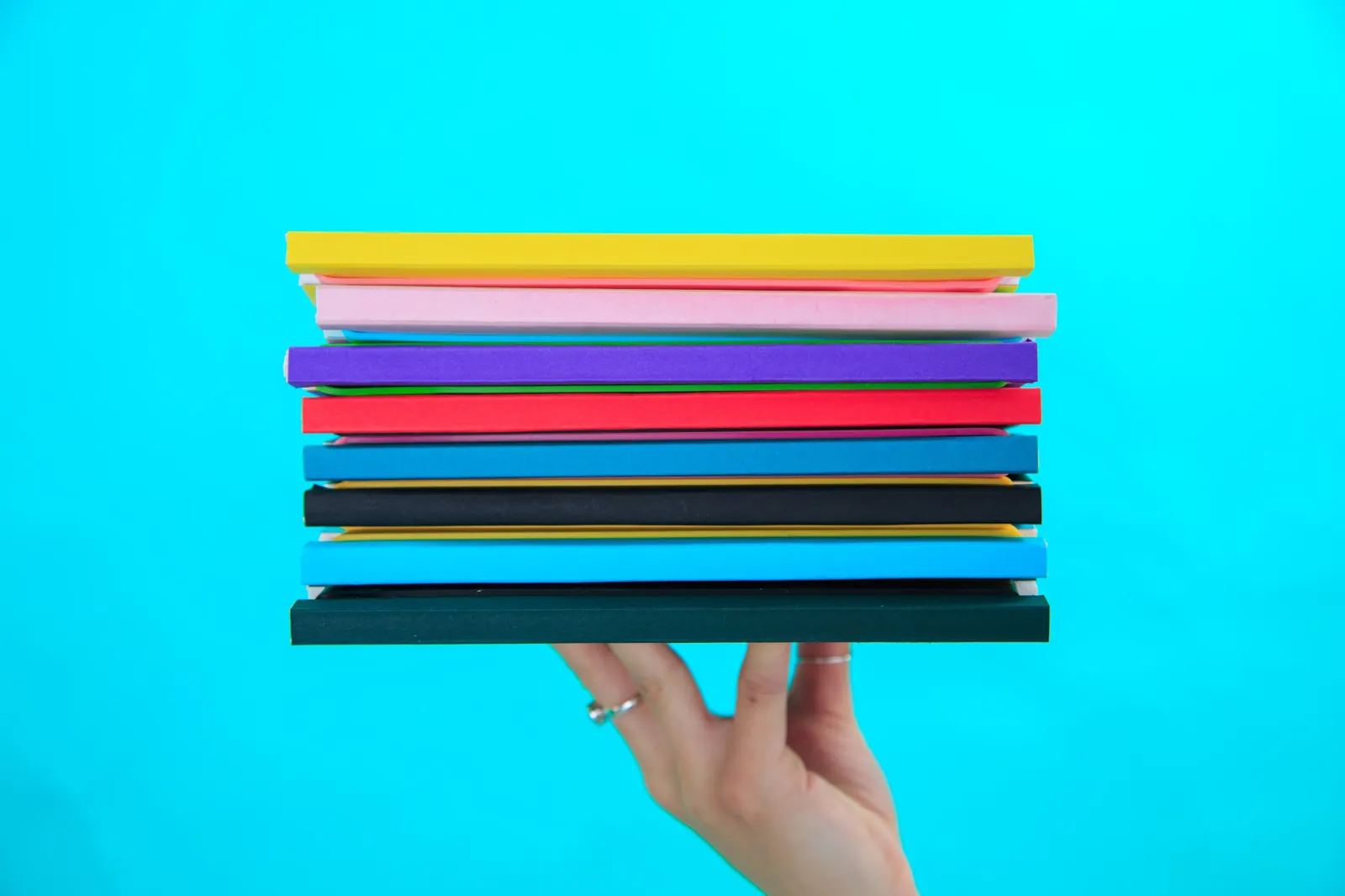 Hand balancing a stack of colourful Book for Life refillable notebooks against a bright cyan background