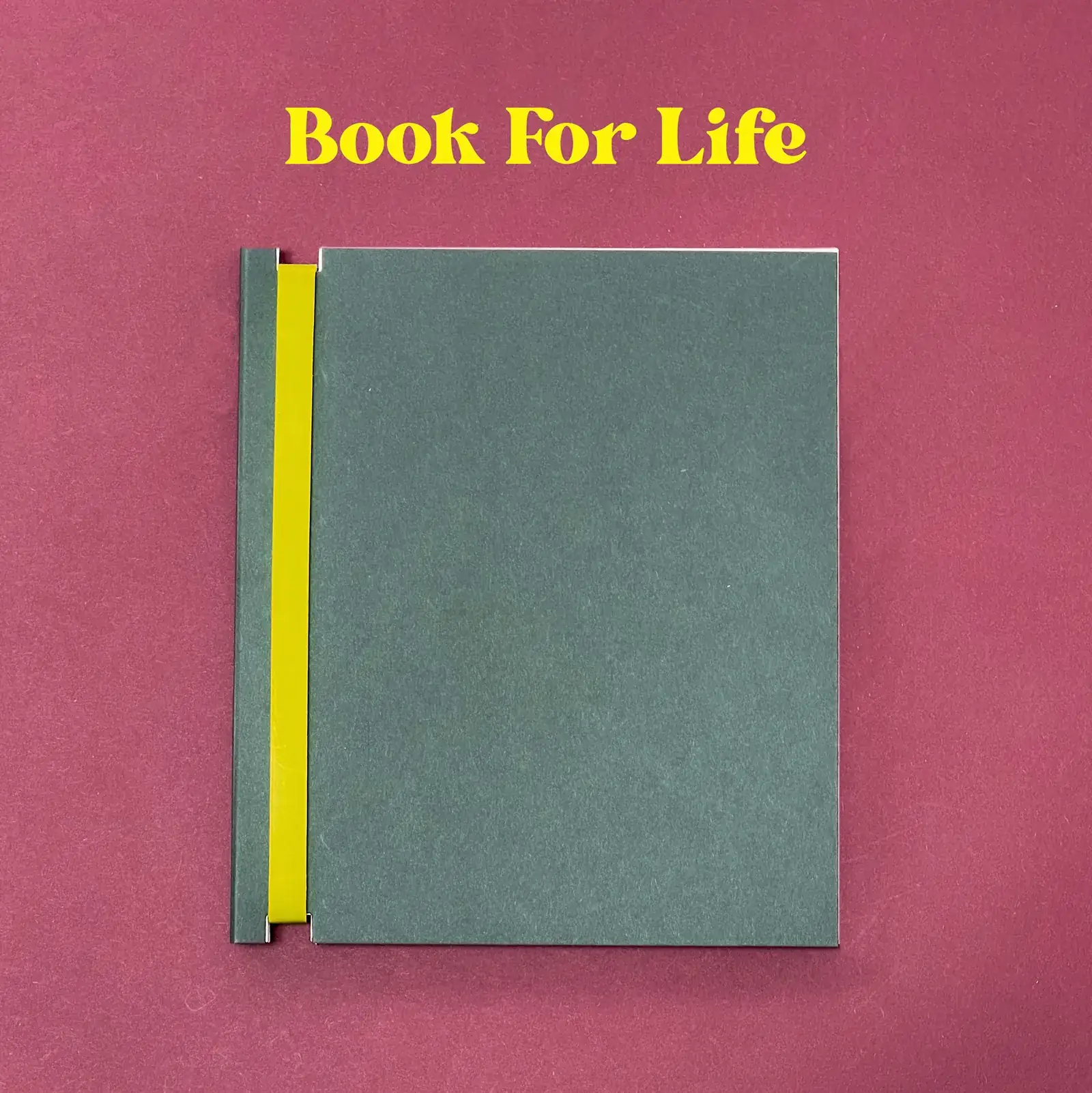 Dizzy Ink Book for Life refillable notebook in forest green with a yellow spine strip, on a burgundy backdrop under yellow Book For Life lettering