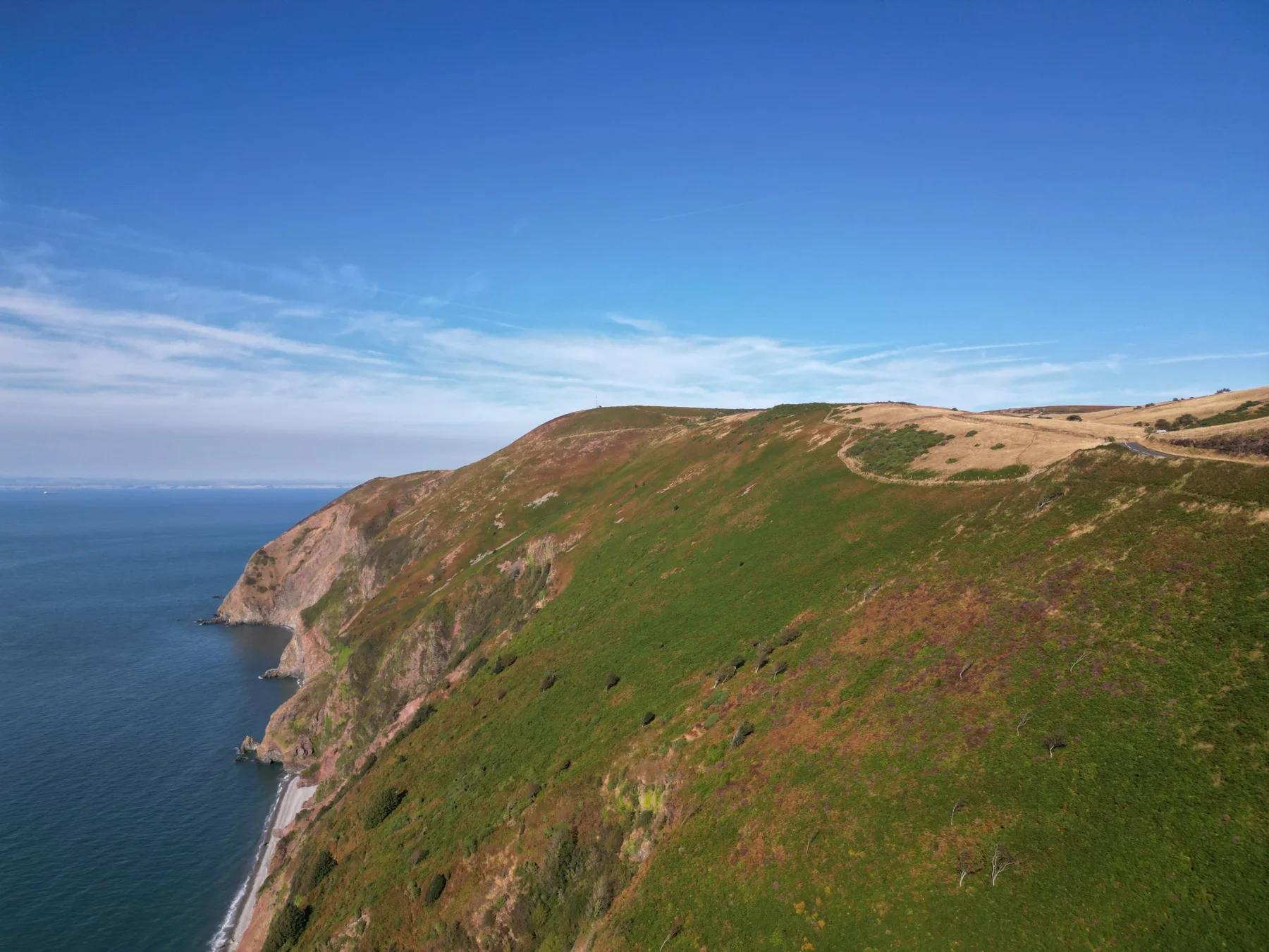 Drone photo of steep green and heather covered cliffs dropping to a shingle beach and the sea on the North Devon coast under a blue sky