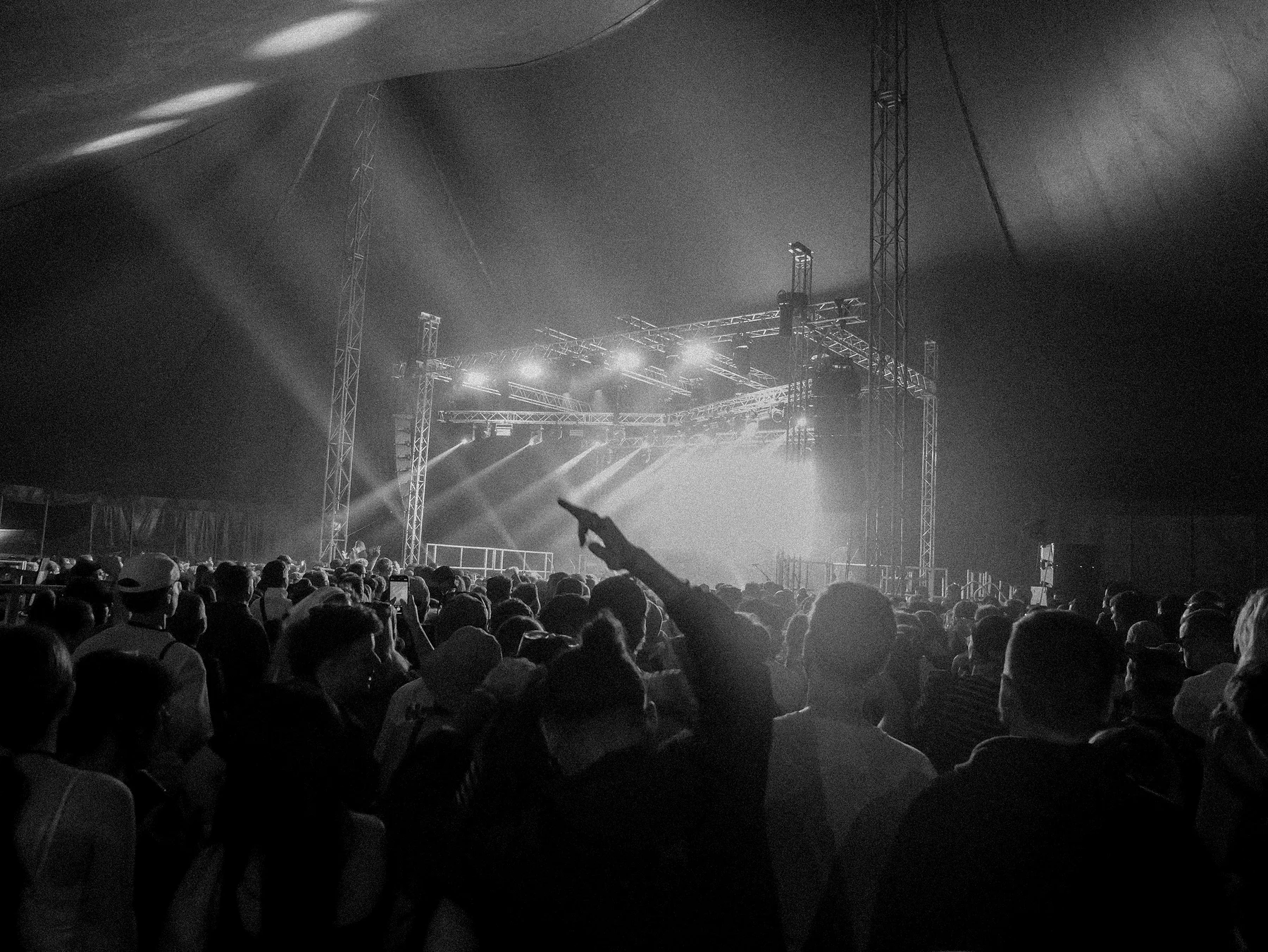 Black and white photograph of a festival crowd facing a big top stage under bright lights, one fan pointing up