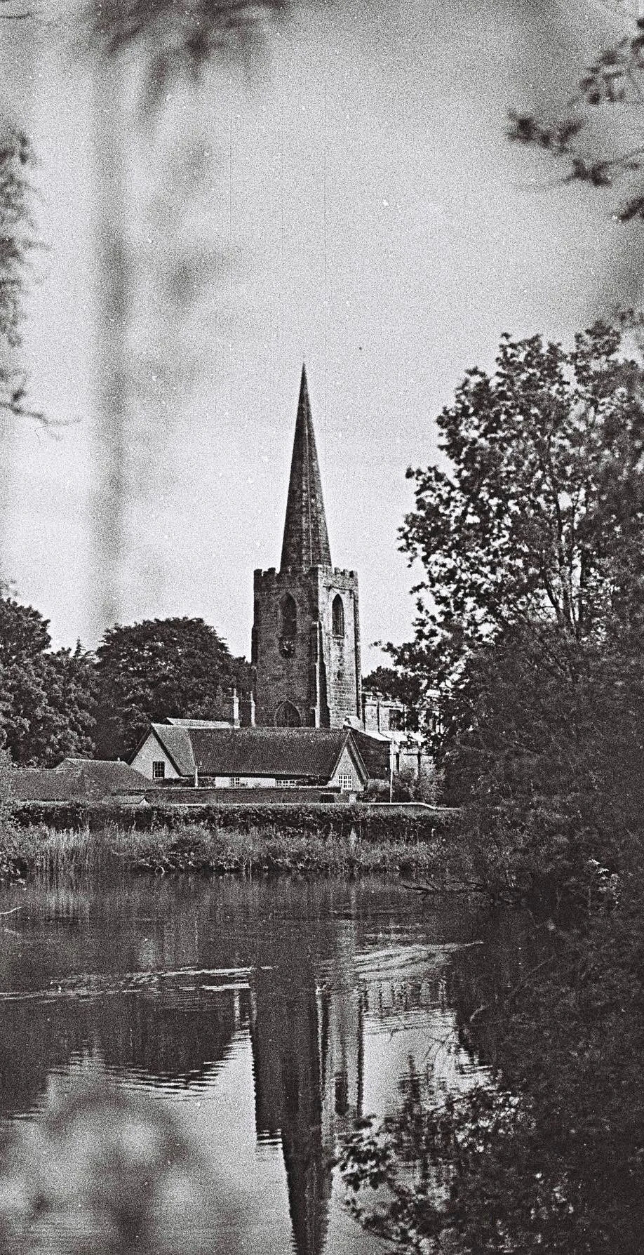 Black and white film photograph of a stone church spire reflected in still water, framed by trees