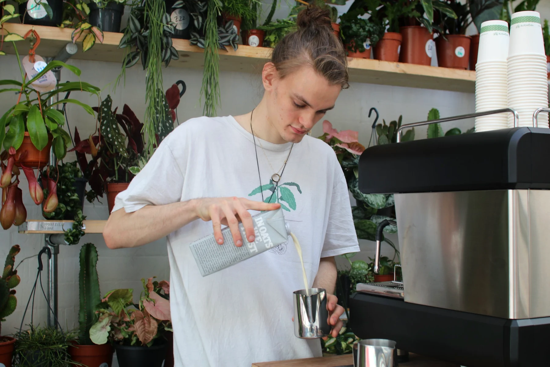 The Lono Espresso barista pouring oat milk into a steel jug beside the espresso machine, shelves of potted plants behind