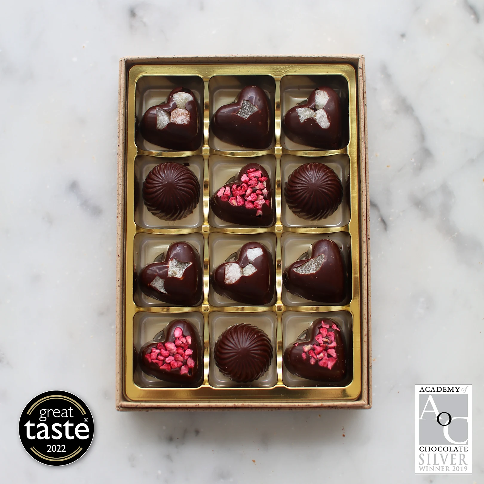 Overhead shot of a box of twelve vegan dark chocolate hearts and bonbons topped with raspberry and crystallised ginger, on white marble, with Great Taste 2022 and Academy of Chocolate Silver awards