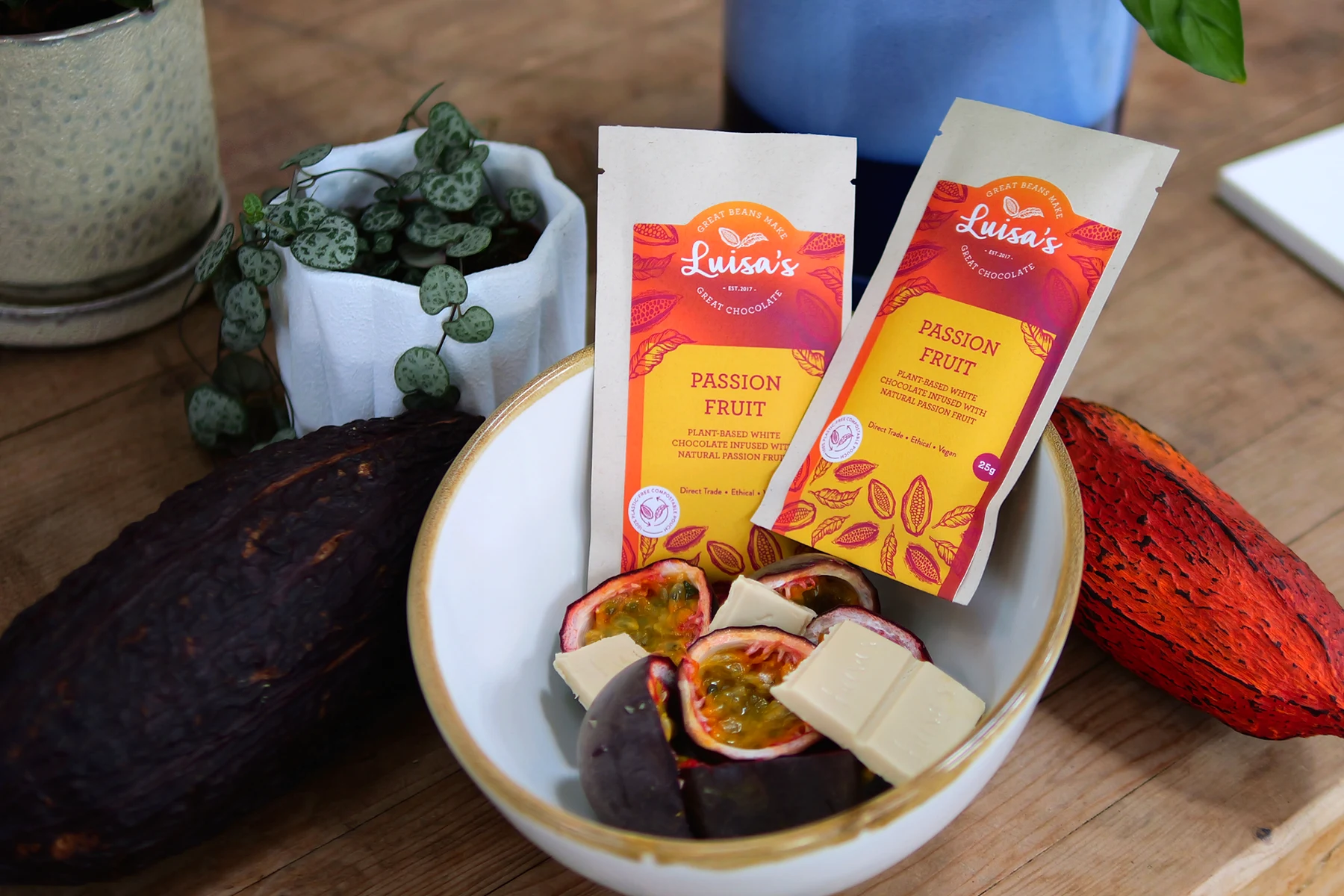 Two Luisa's Passion Fruit plant-based white chocolate pouches in a bowl of halved passion fruit and white chocolate squares, between two cacao pods