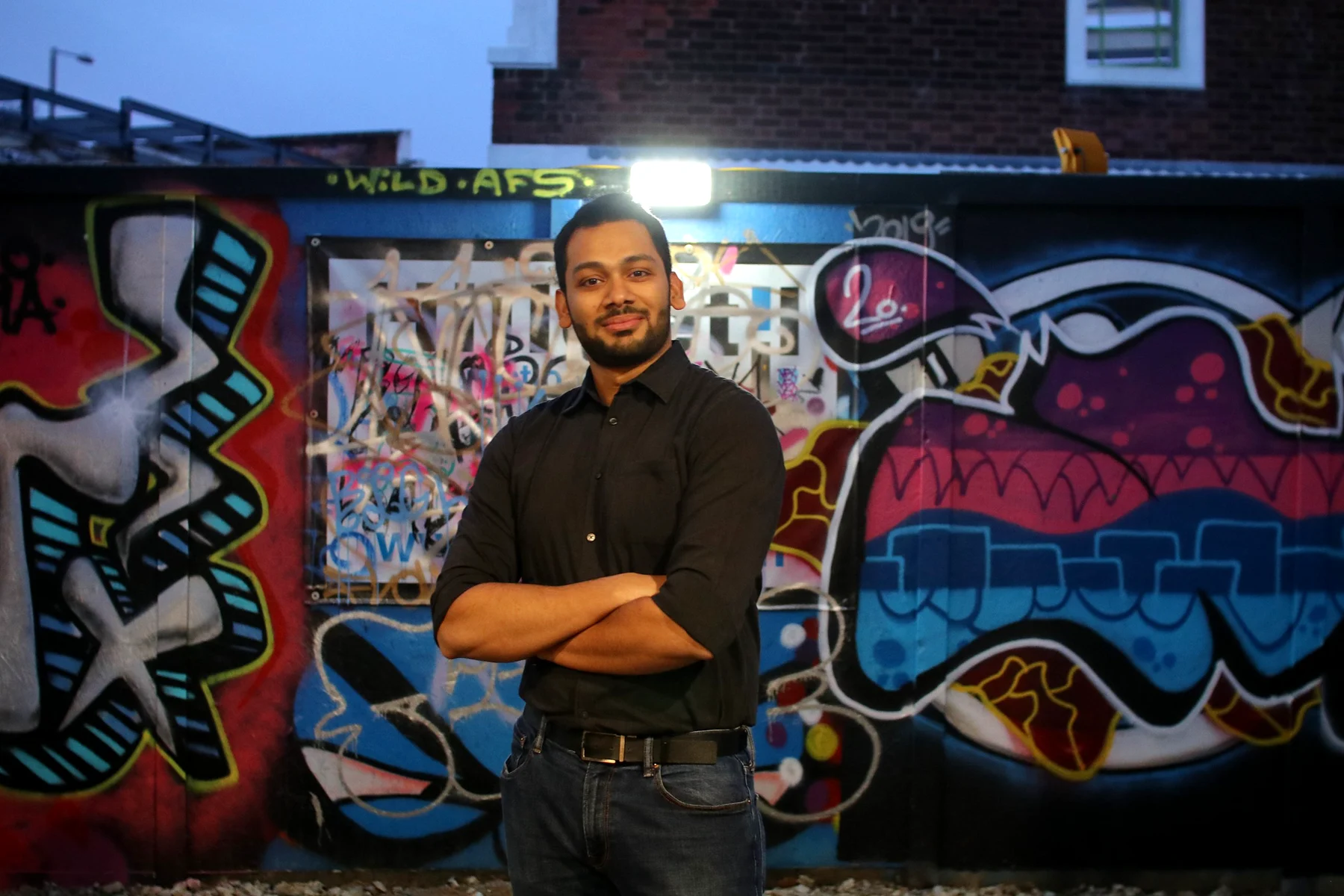 A man in a black shirt with arms folded in front of a bright graffiti wall at dusk, lit by a floodlight behind him