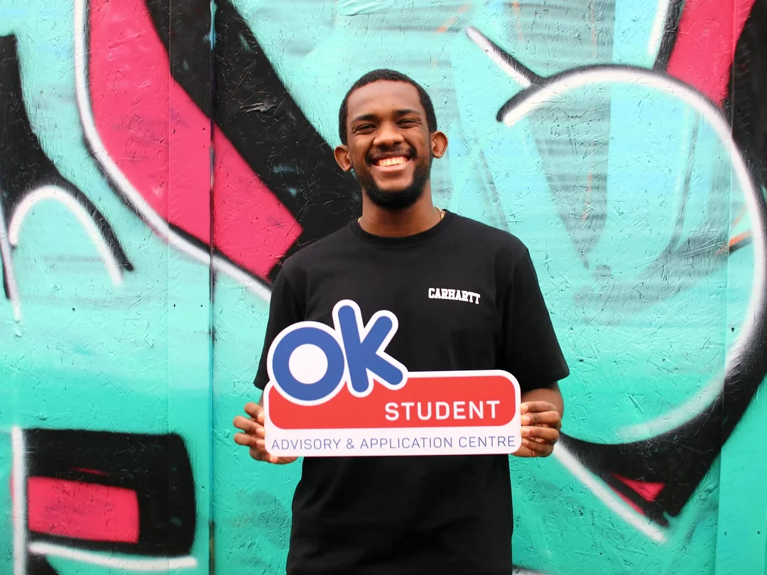 Smiling student holding an OK Student sign in front of a teal and pink graffiti wall