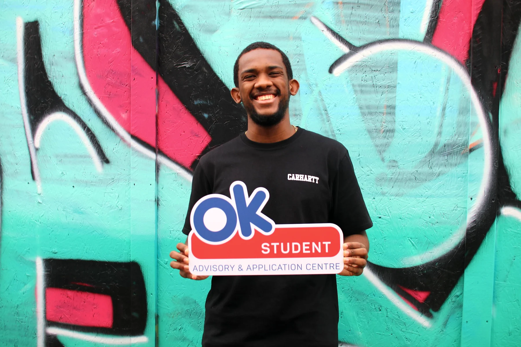 A smiling student in a black t-shirt holding an OK Student sign in front of a teal, pink and black graffiti wall