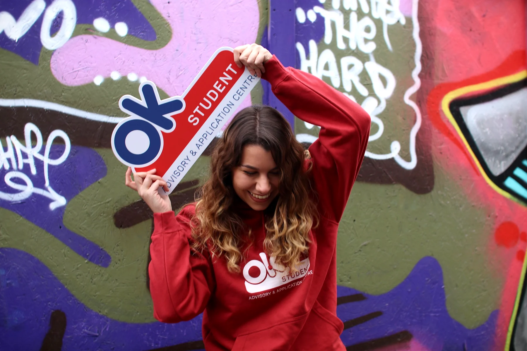 A laughing student in a red OK Student hoodie holding a key shaped OK Student sign over her head against a graffiti wall