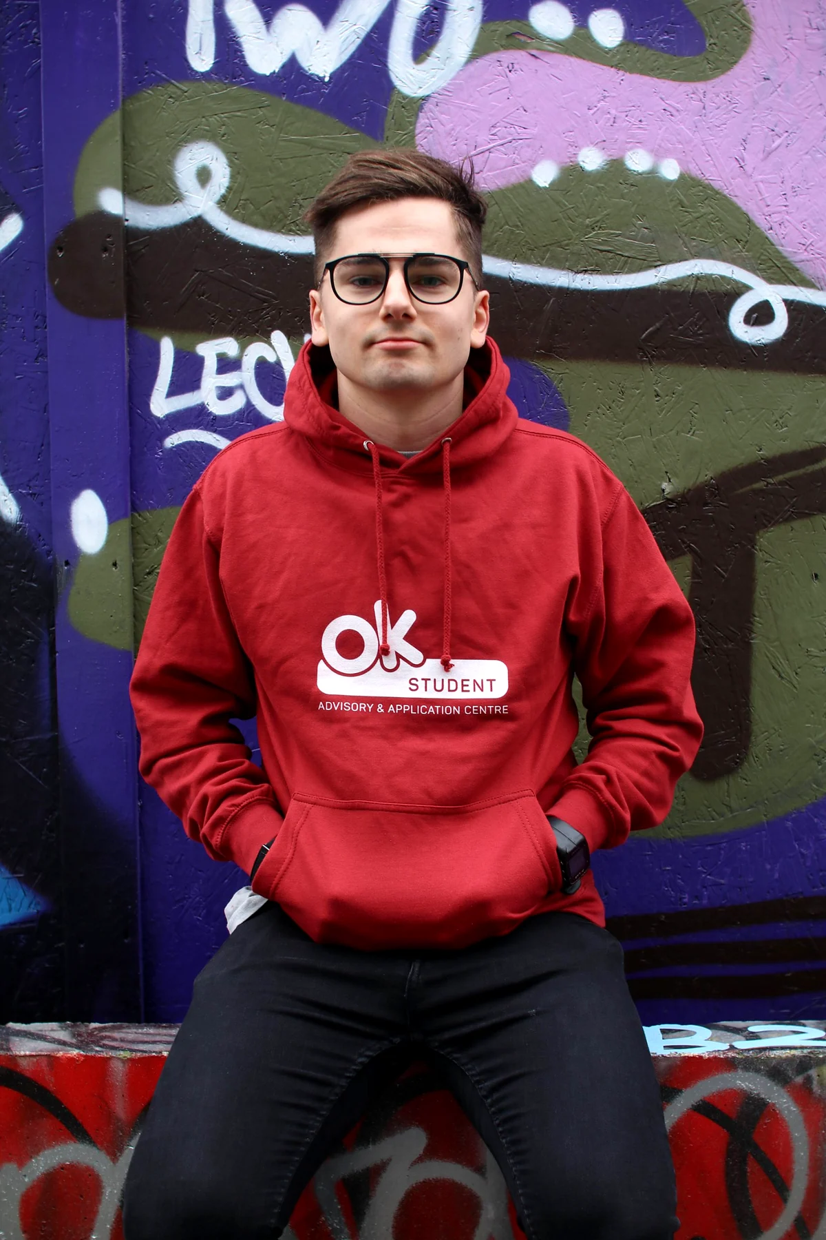 A student in a red OK Student hoodie and glasses sitting in front of a purple, green and pink graffiti wall