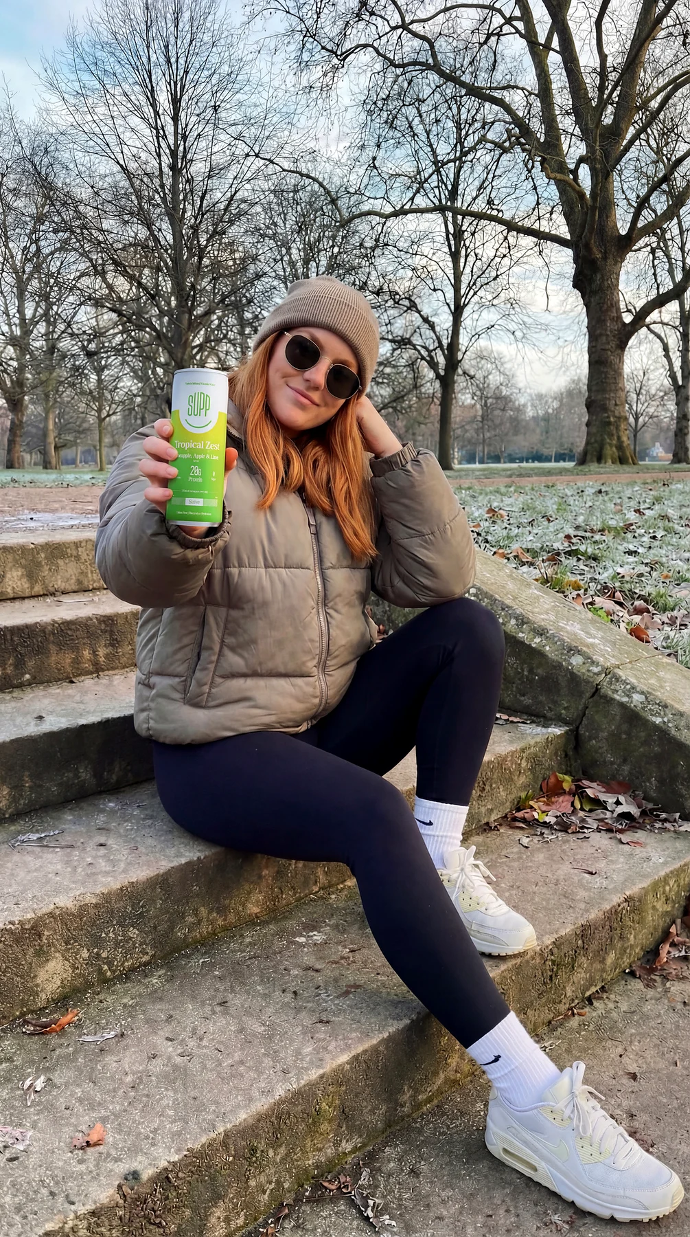 A young woman in a puffer jacket sitting on park steps in winter, smiling and holding up a can of Supp