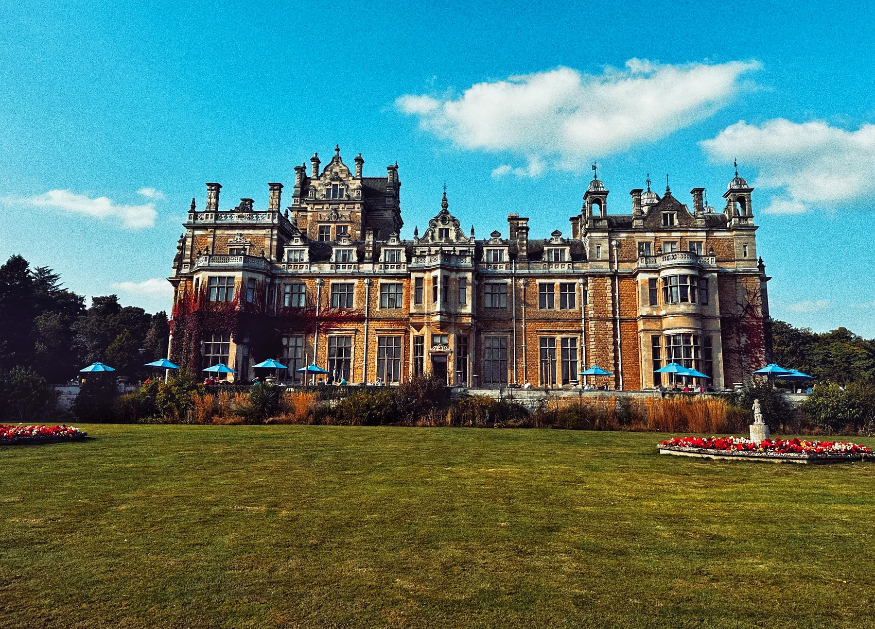 A grand Victorian country house with towers and chimneys behind a wide green lawn under a blue sky