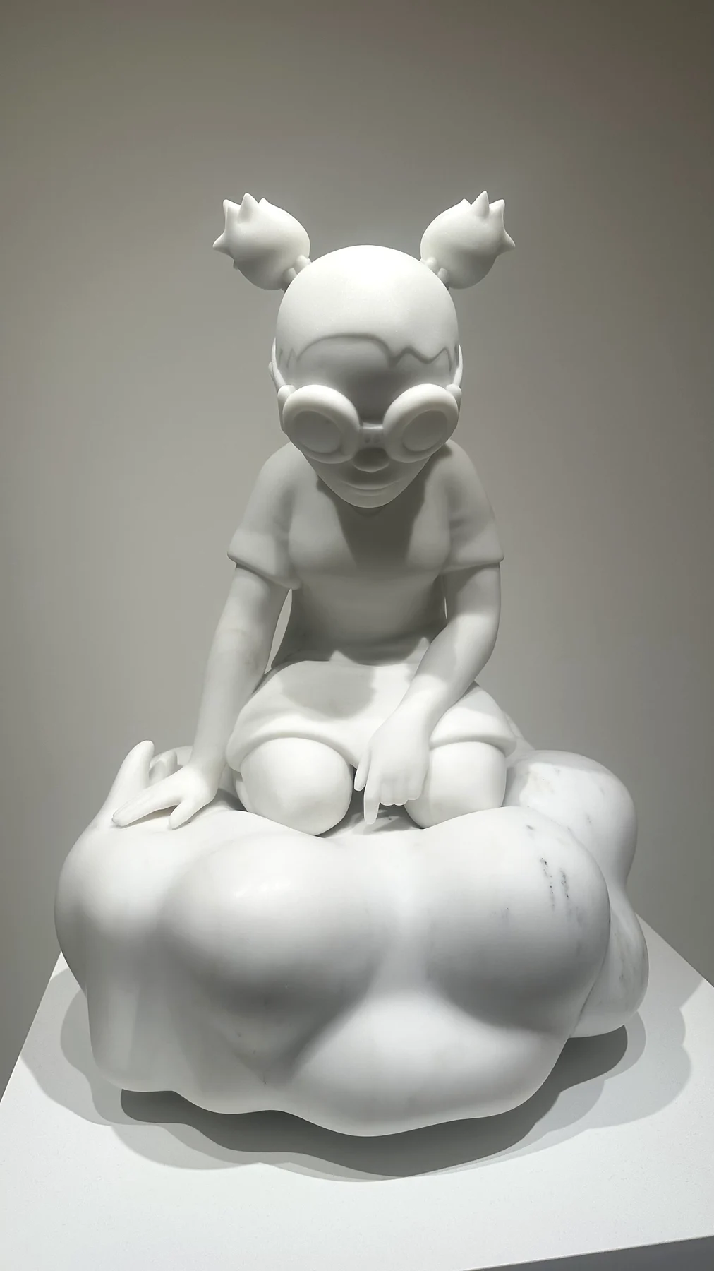 A white sculpture of a cartoon-like figure with goggles sitting on a cloud, in a gallery