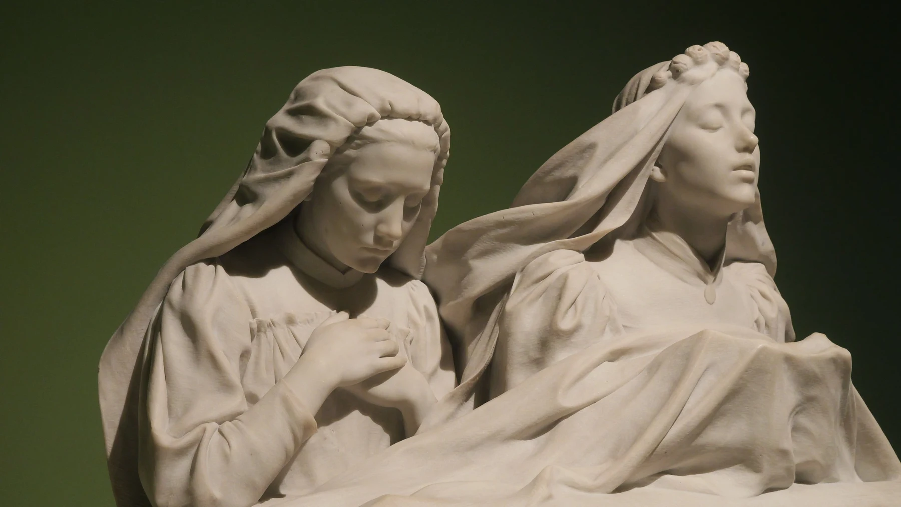 A white marble sculpture of two veiled young women, one praying with eyes down, the other with eyes closed, lit against a dark green wall