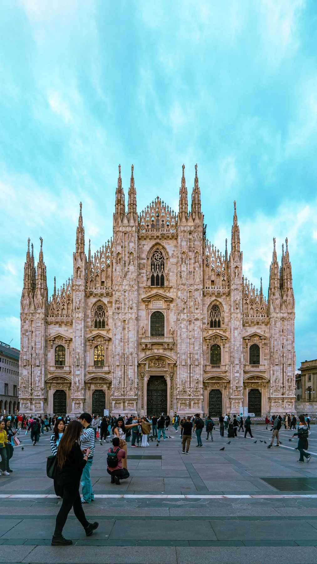 The marble facade and spires of Milan Cathedral, the Duomo di Milano, under a turquoise sky with people crossing the piazza