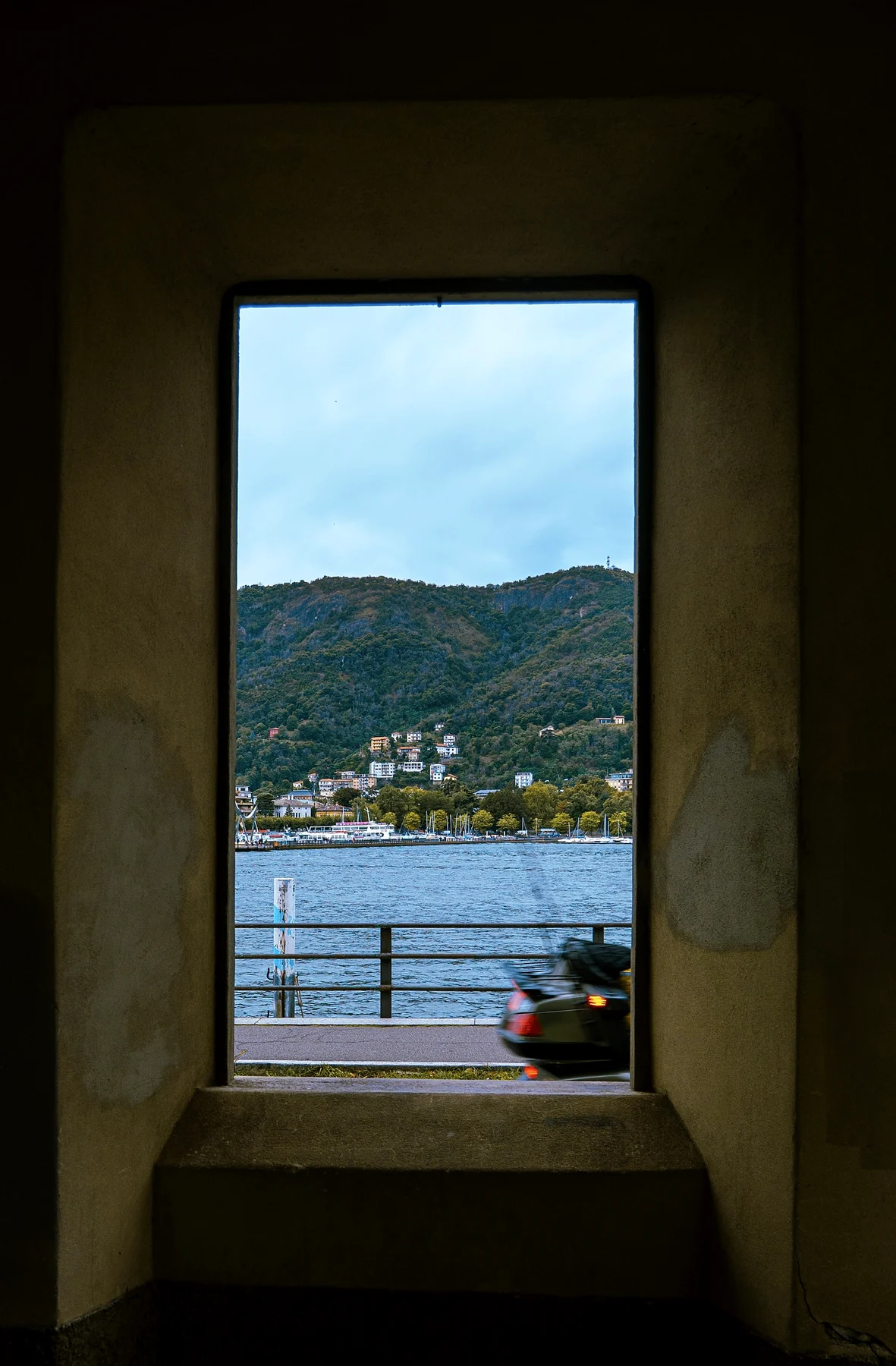 A deep stone window framing a lake, a waterfront promenade and wooded mountains beyond