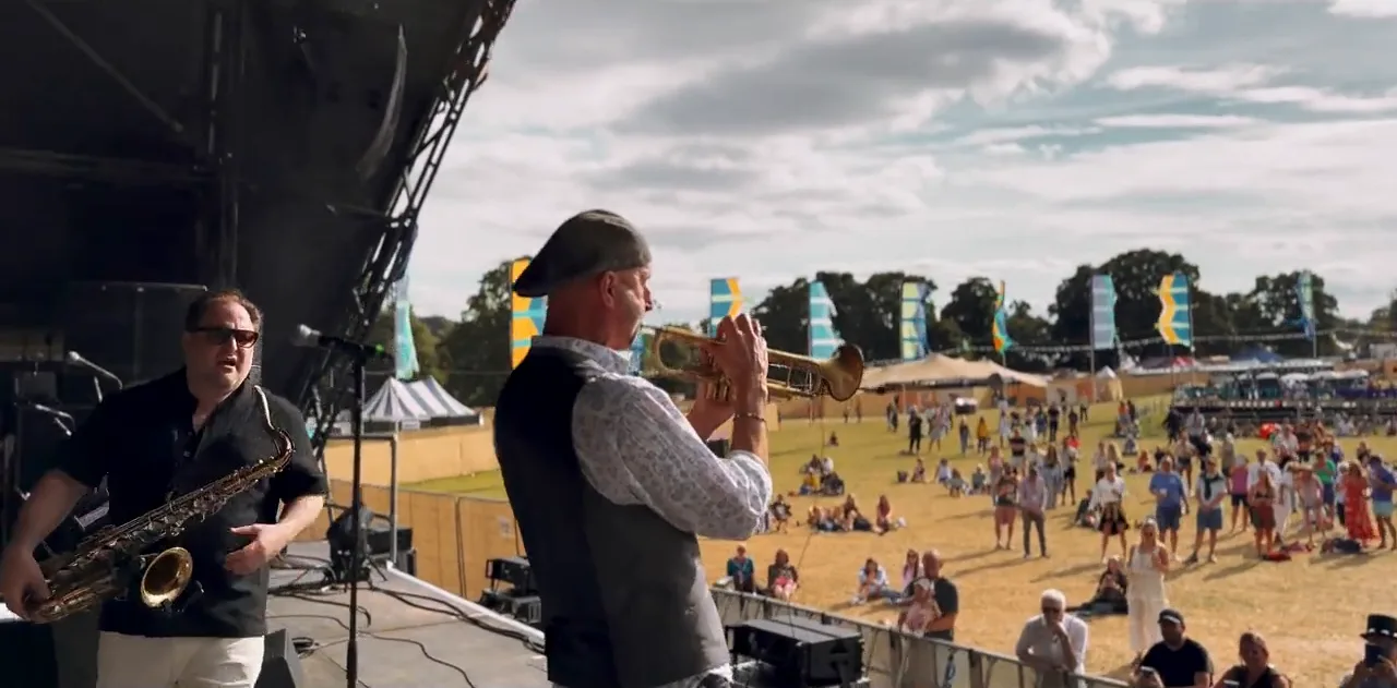 Saxophone and trumpet players on a festival stage with the crowd and tents behind
