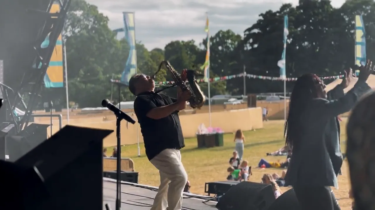 A saxophonist leaning back mid solo beside a singer with her arm raised, festival flags behind