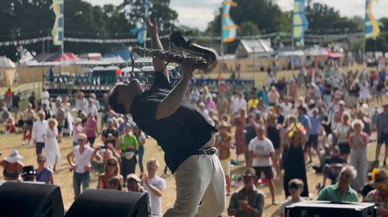 A saxophonist bending backwards mid solo at the front of the stage, a big festival crowd cheering below