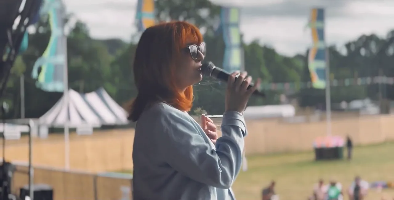 A singer with red hair and sunglasses singing into a microphone on a festival stage