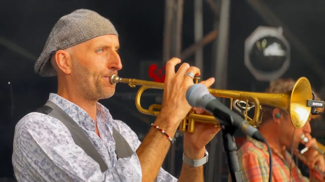 A trumpet player in a grey flat cap playing into the mic, close up